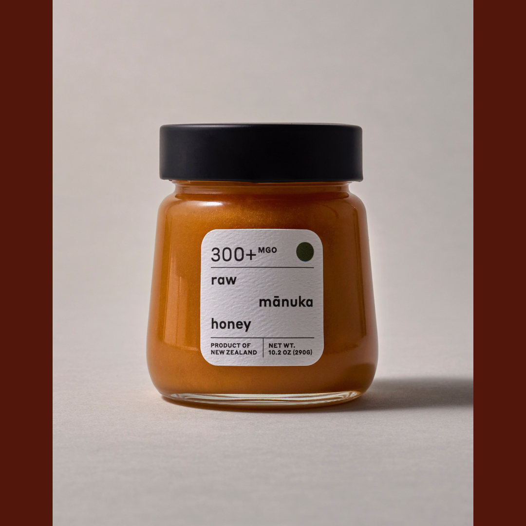 A glass jar of raw mānuka honey with a black lid and a white label, set against a neutral light gray background with dark brown vertical bars on the sides.