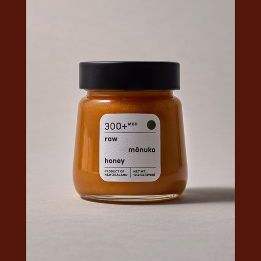 A glass jar of raw mānuka honey with a black lid and a white label, set against a neutral light gray background with dark brown vertical bars on the sides.