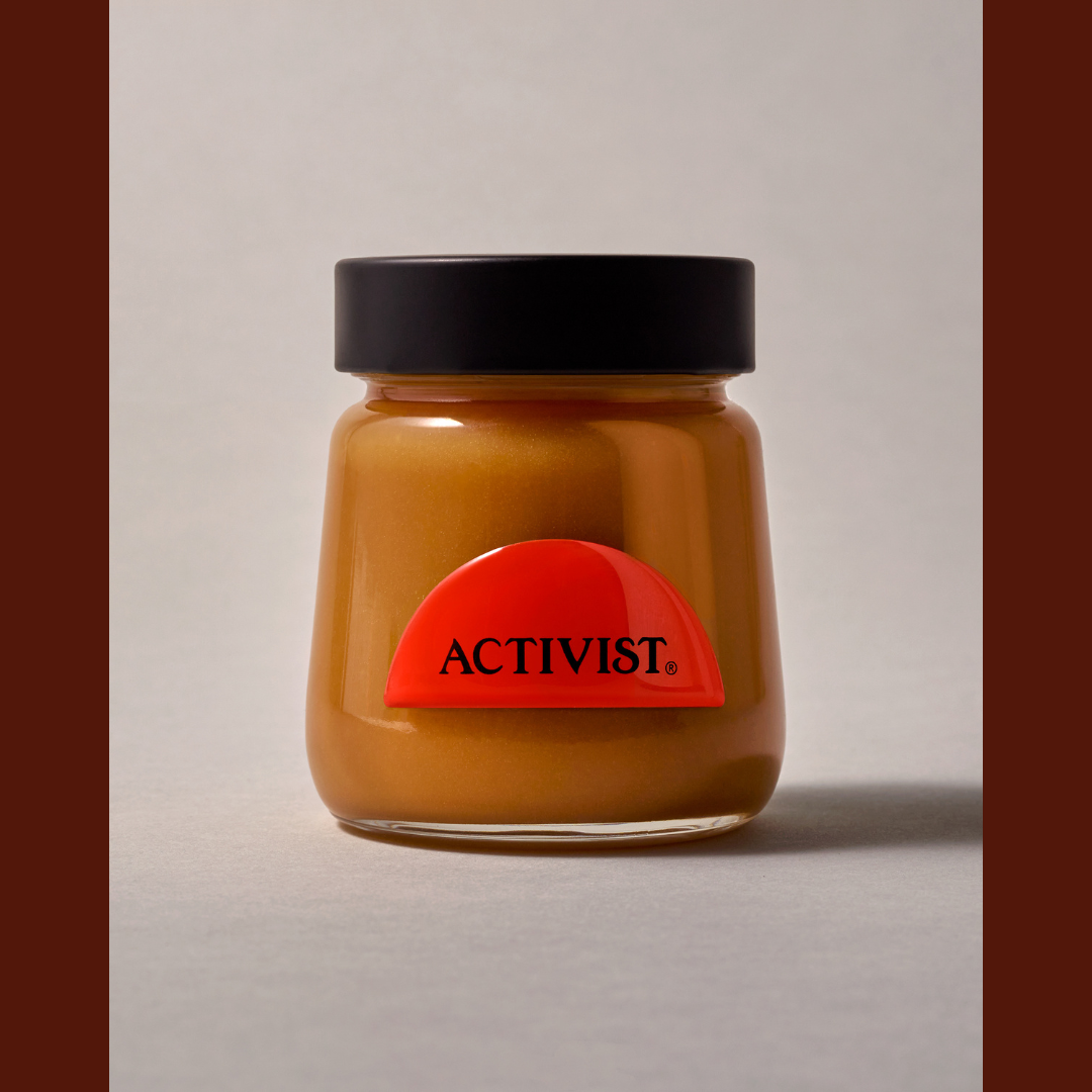 A glass jar of ACTIVIST. brand spread with a black lid and a red semi-circular label, set against a neutral gray background with dark brown vertical bars on the sides.