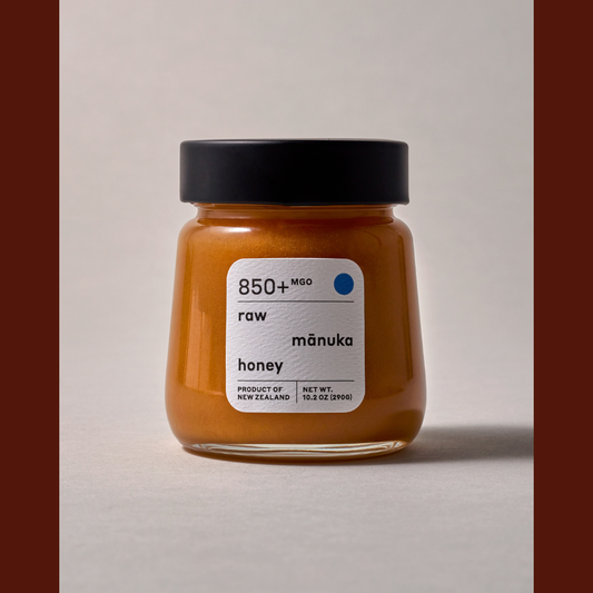 A glass jar of raw mānuka honey with a black lid and a white label, set against a neutral light gray background with dark brown side borders.